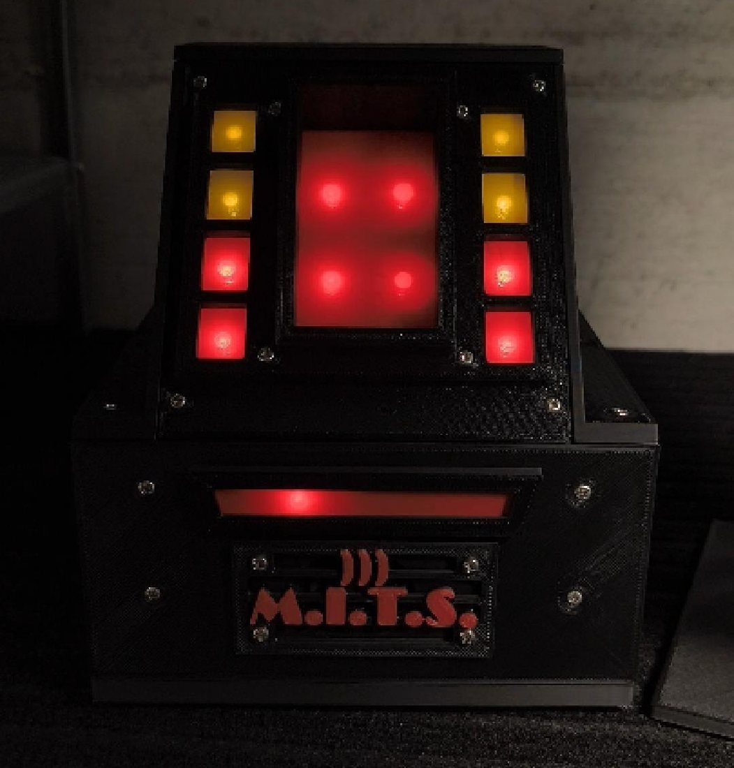 M.I.T.S., dispositivo stampato in 3D nero, con una griglia di LED rossi accesi al centro, pulsanti gialli e rossi ai lati e la scritta M.I.T.S. sul frontale.